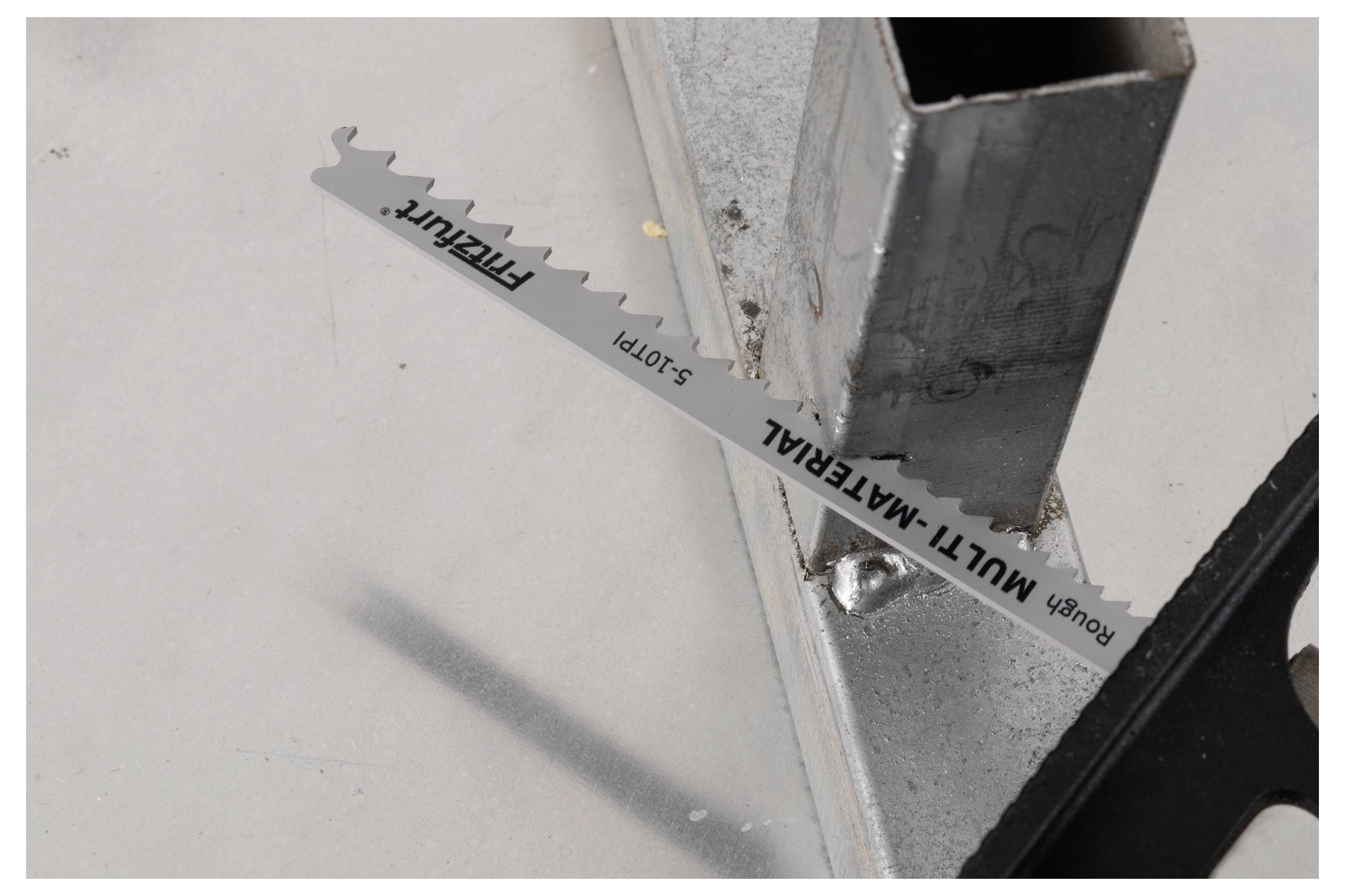 Eine Stichsägeblatt aus Metall, beschriftet mit "MULTI-MATERIAL" und "ROUGH", wird in eine Stichsäge eingesetzt, die auf einer metallischen Oberfläche neben einem Stahlrahmen ruht.