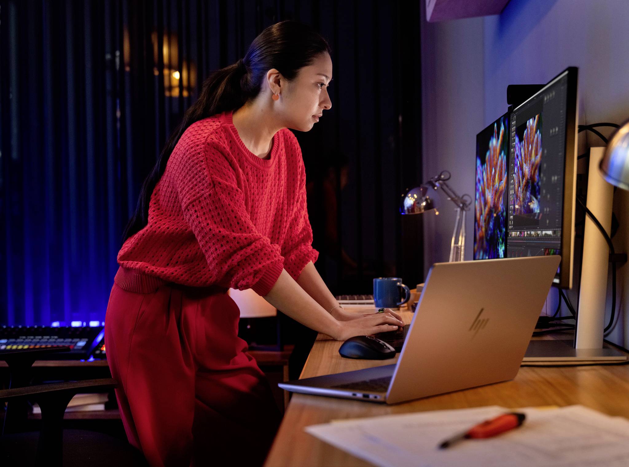 Eine Person in einem roten Pullover arbeitet konzentriert an einem Computer mit mehreren Monitoren, in einem Büro bei schwacher Beleuchtung.