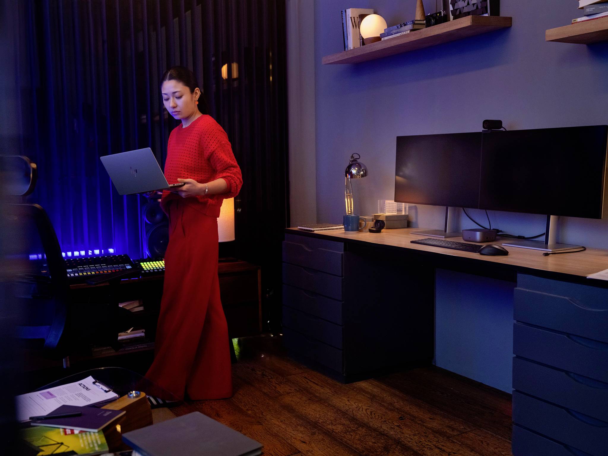 Eine Frau in einem roten Outfit steht in einem modernen Büro mit Computern und blauer Beleuchtung, während sie auf einen Laptop schaut.