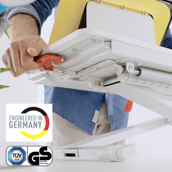 'Person justiert weißen Schreibtischständer mit rotem Hebel.' Aufdruck 'Engineered in Germany', TÜV- und GS-Zertifikate sichtbar.
