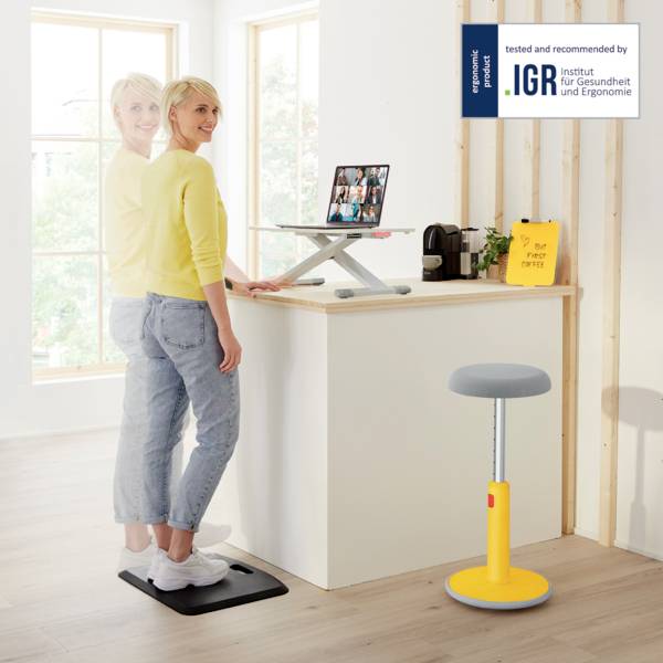 Zwei Frauen stehen in einem hellen Raum. Eine arbeitet an einem Stehtisch mit Laptop, während die andere auf einer Gleichgewichtsmatte steht.