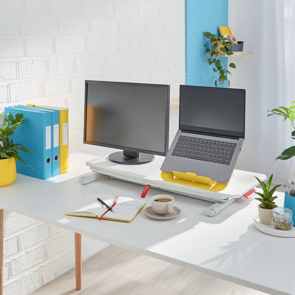 Moderner Schreibtisch mit Laptop und Monitor, umgeben von Pflanzen. Nebenan stehen farbige Ordner und Schreibutensilien.