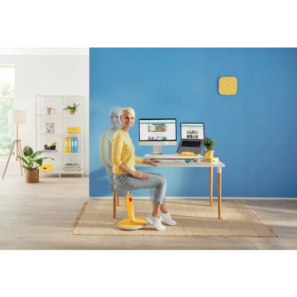Frau sitzt an einem Schreibtisch mit zwei Bildschirmen, auf denen Websites zu sehen sind. Moderner, bunter Raum mit gelbem Hocker und blauer Wand.