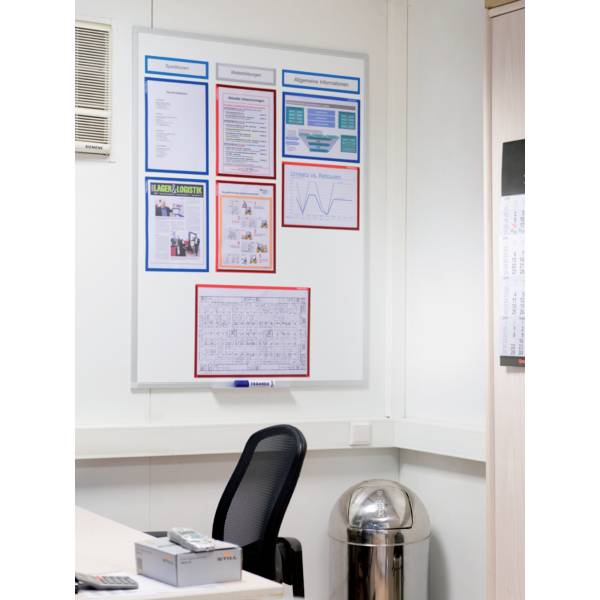 Ein Whiteboard im Büro mit Informationsblättern und Diagrammen hängt an der Wand über einem Schreibtisch mit Stuhl und Abfalleimer.