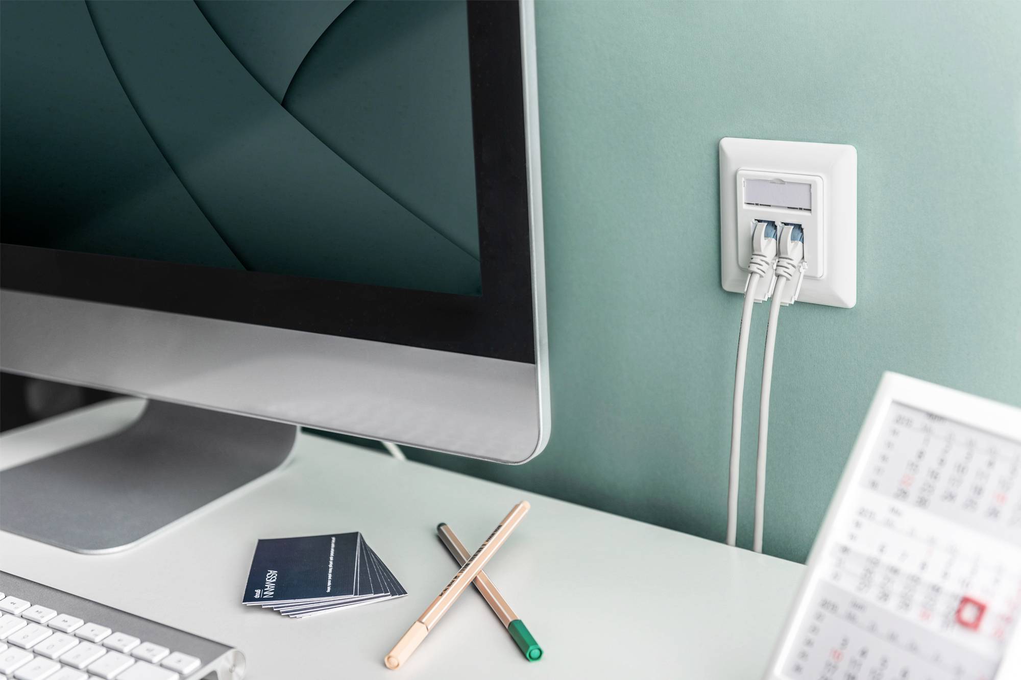 Arbeitsbereich mit Computer, Steckdose mit eingesteckten Kabeln, und zwei Stiften auf dem Schreibtisch neben Visitenkarten.