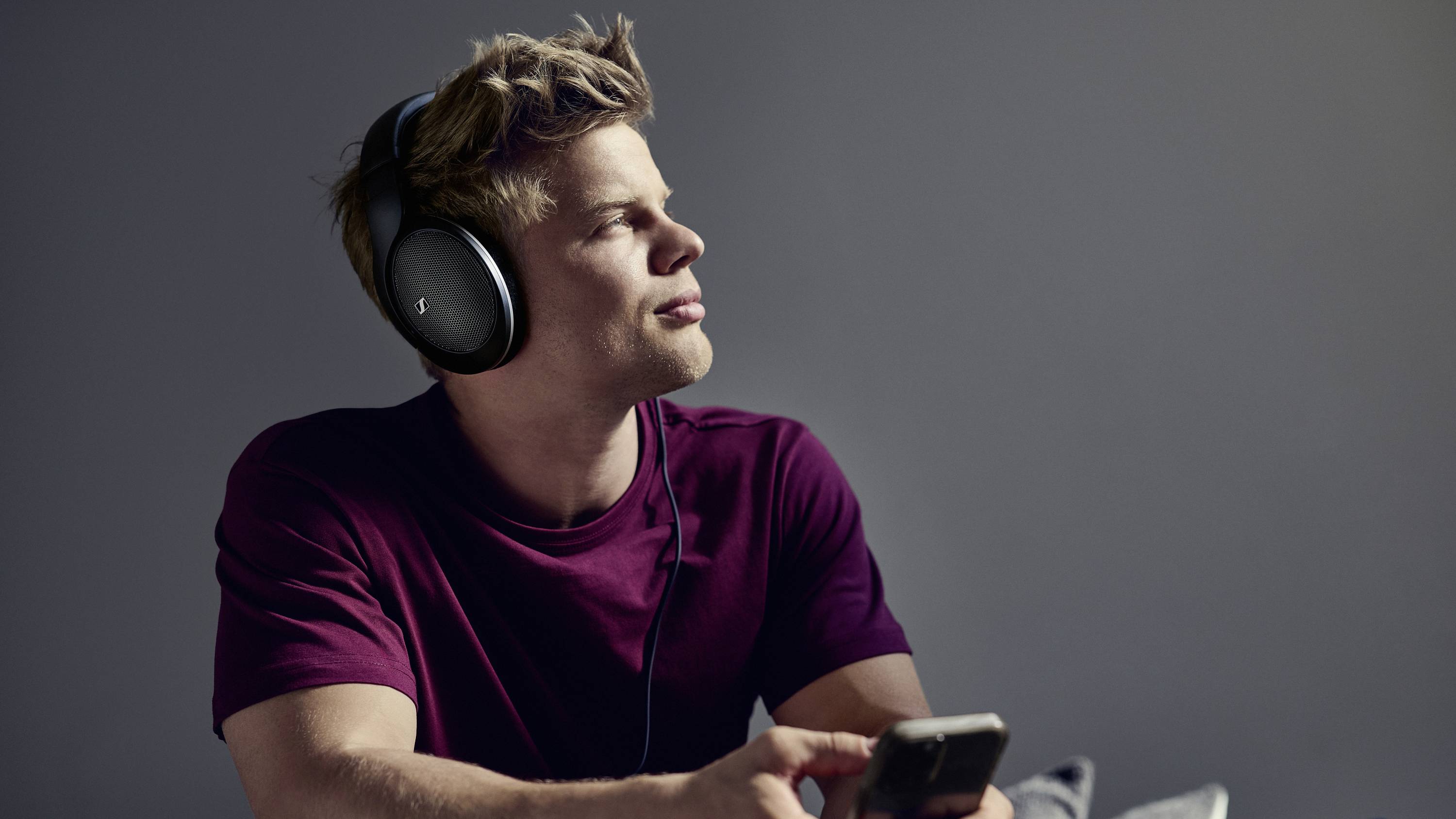 Ein Mann mit Kopfhörern sitzt entspannt und schaut nachdenklich zur Seite. In der Hand hält er ein Smartphone.