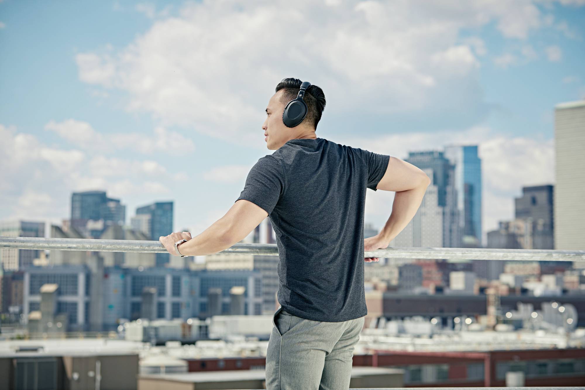 Eine Person mit Kopfhörern schaut von einer städtischen Dachterrasse in die Ferne. Im Hintergrund sind Wolken und Hochhäuser zu sehen.