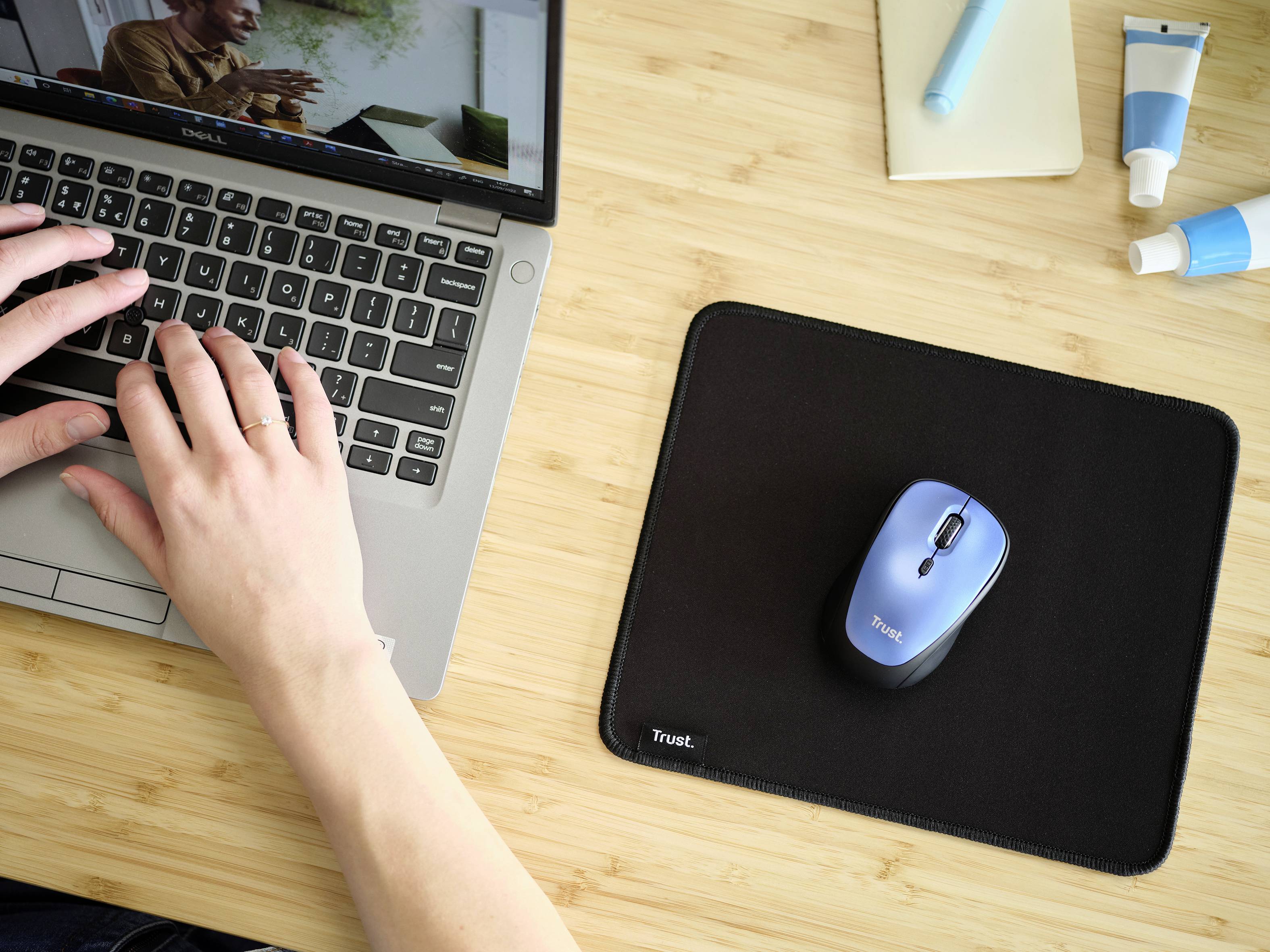 Person tippt auf Laptop, daneben eine Maus auf einem Mauspad. Tisch hat Pflegeprodukte und Notizheft. Arbeitsplatz-Szene.