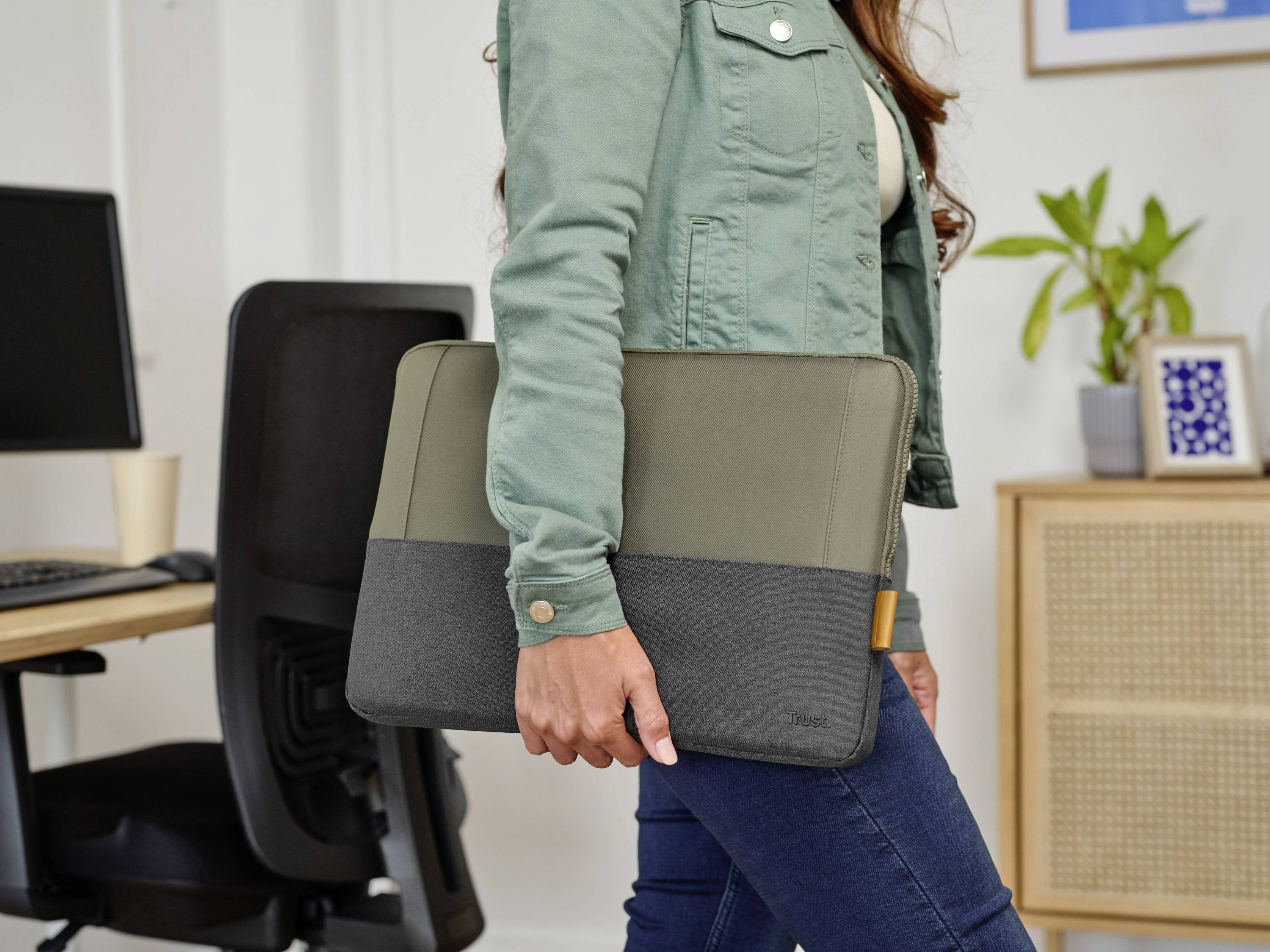 Eine Person hält eine zweifarbige Laptop-Tasche in einem Büro. Im Hintergrund sind ein Schreibtischstuhl und ein Schrank mit Pflanze zu sehen.