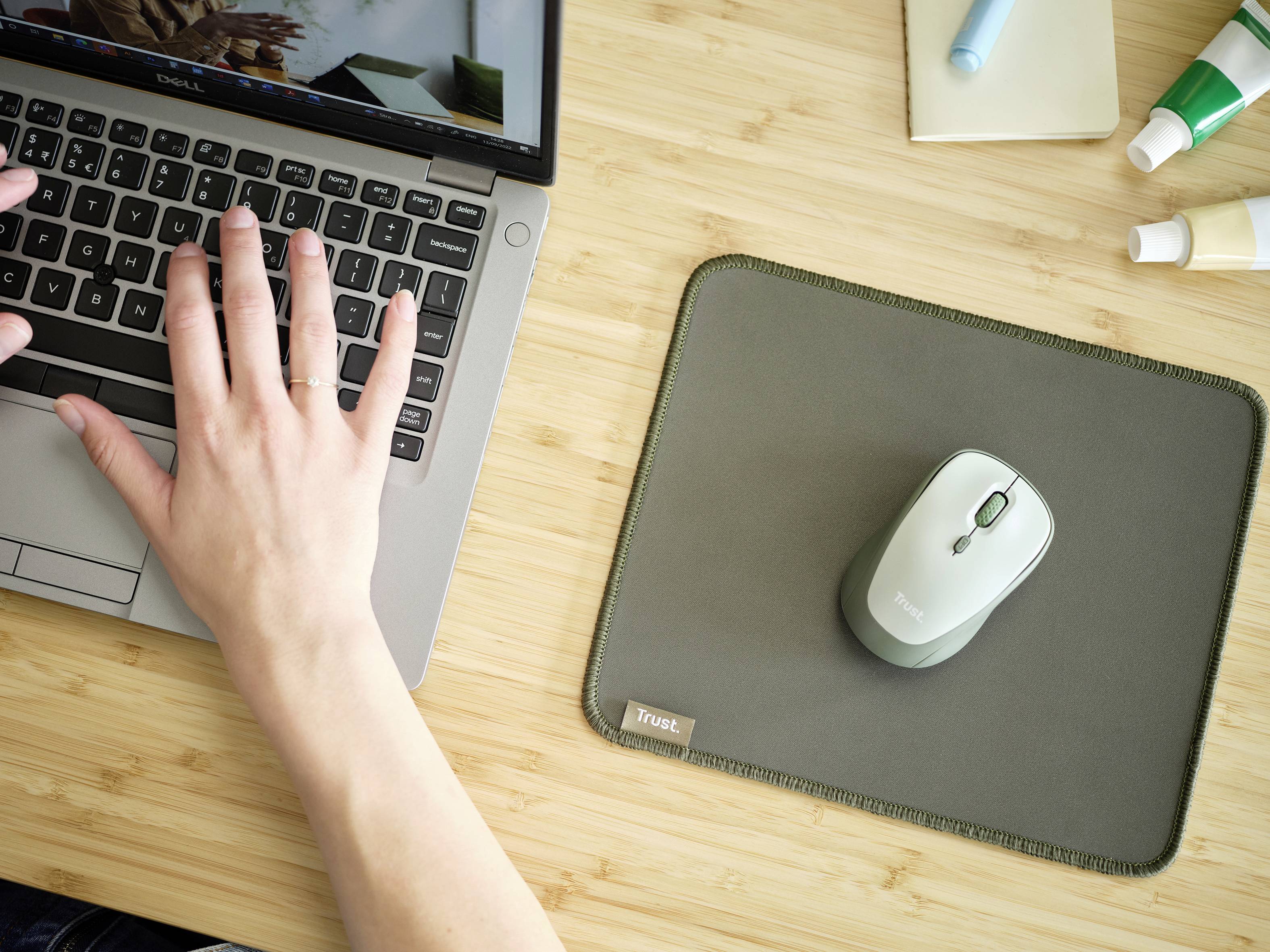 Eine Hand tippt auf einem Laptop, daneben eine Computermaus auf einem Mauspads auf einem Holztisch, umgeben von Büroartikeln.