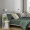 Gemütliches Schlafzimmer mit modernem grauem Bett, grünen Bettdecken und Kissen. Nachttisch mit Pflanzen und Lampe, Tageslicht durch das Fenster.