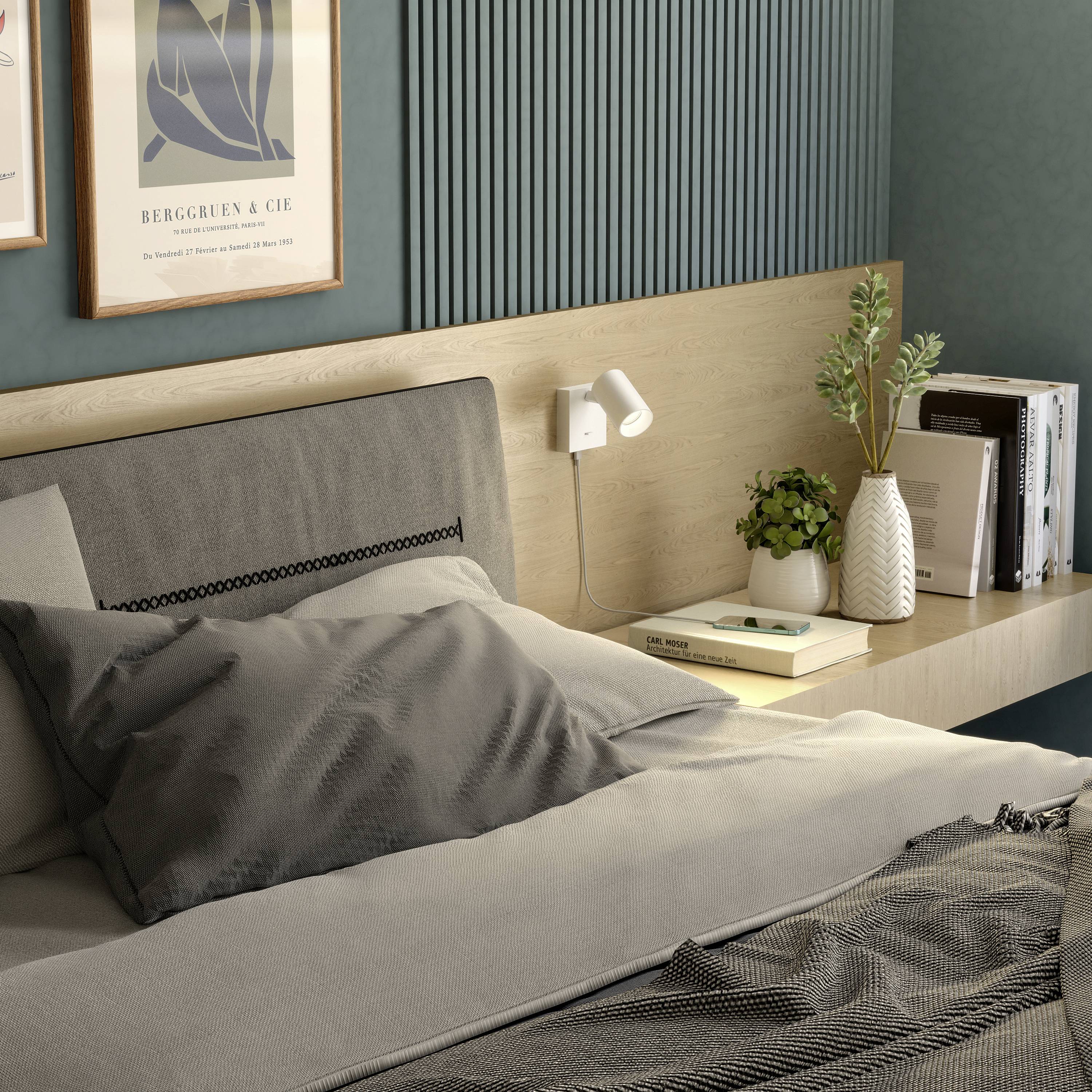Ein gemütliches Schlafzimmer mit einem grauen Bett, einem Nachttisch, Pflanzen, Büchern und Kunst an der Wand.
