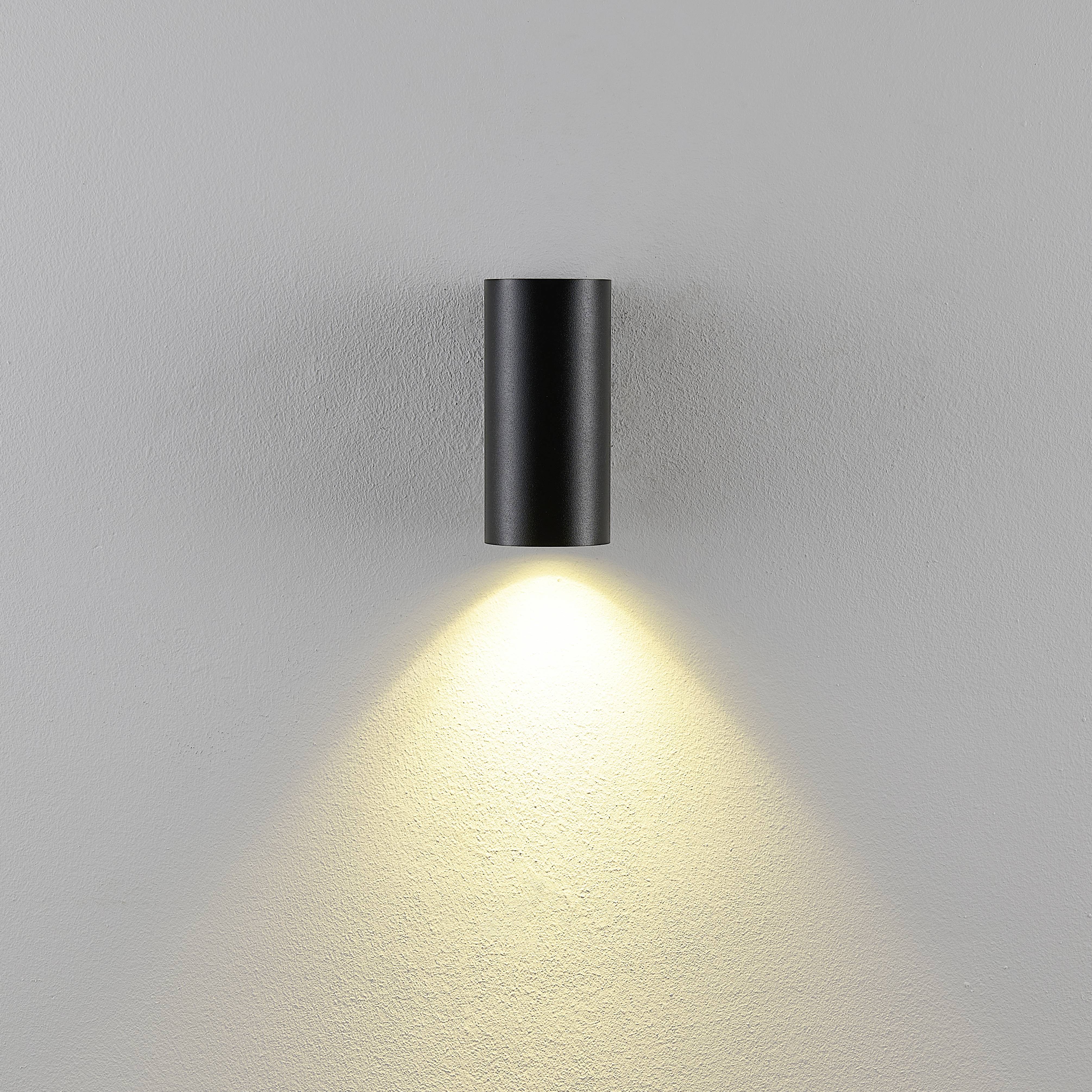 Eine schwarze Wandleuchte strahlt warmes Licht nach unten auf die weiße Wand, wodurch eine angenehme Atmosphäre erzeugt wird.