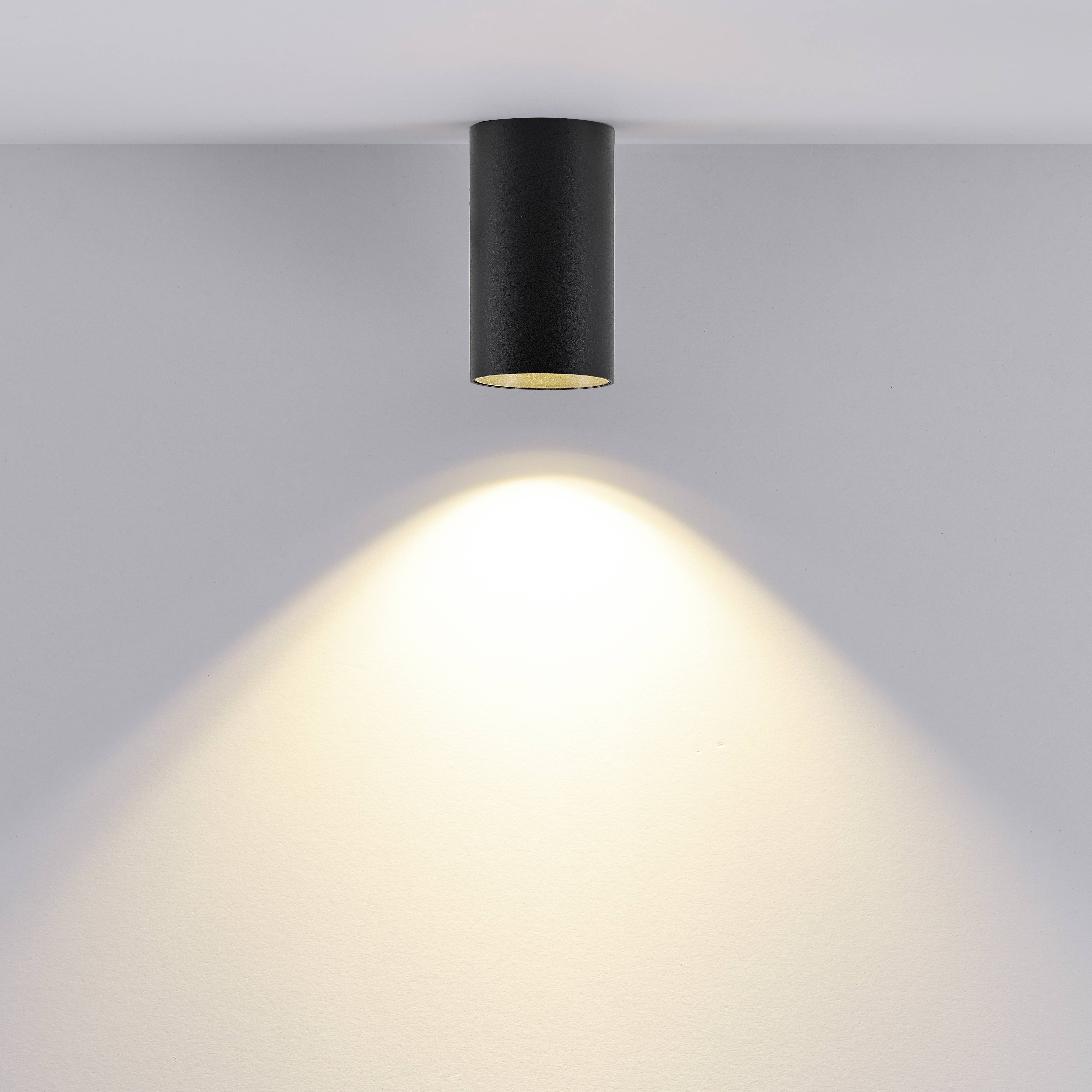 Schwarze Deckenlampe strahlt warmes Licht auf weiße Wand. Minimalistisches Design, Betonung auf Beleuchtungseffekt.
