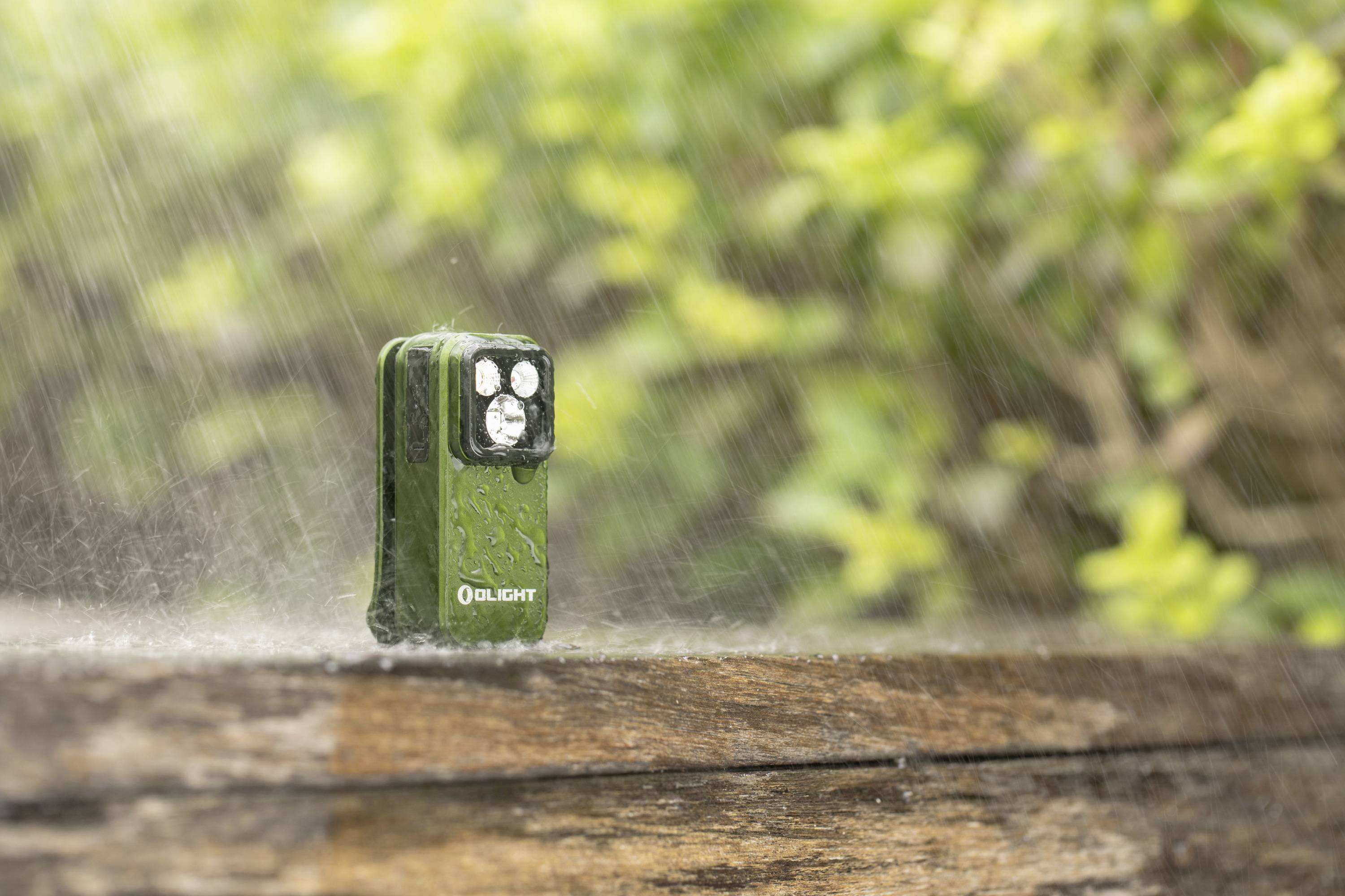 Eine grüne, wasserdichte Taschenlampe steht im Regen auf einem Holzbrett. Im Hintergrund sind unscharfe, grüne Blätter zu sehen.