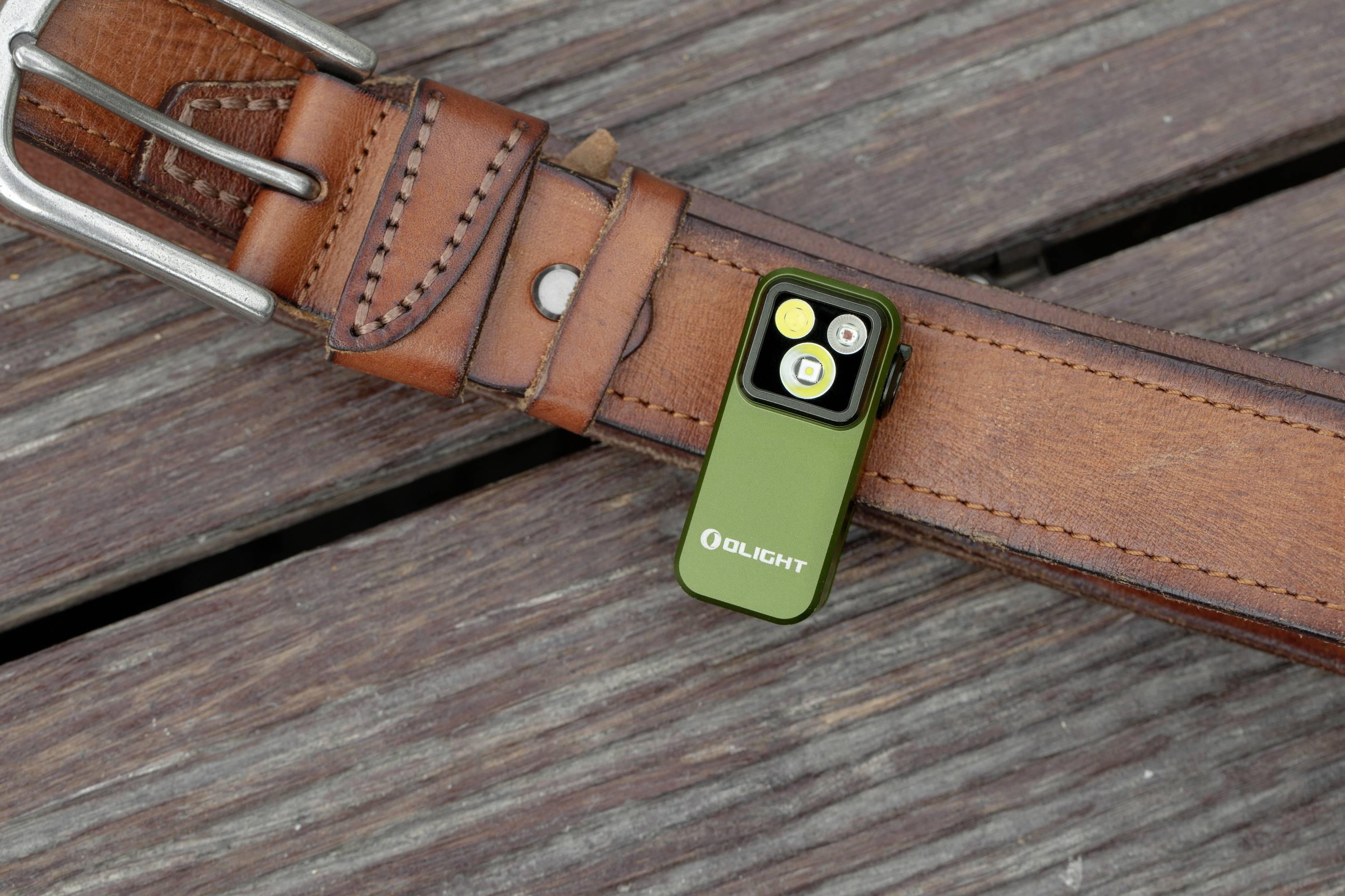 Kleine, grüne Taschenlampe mit Clip an einem braunen Ledergürtel befestigt, auf Holzhintergrund liegend.