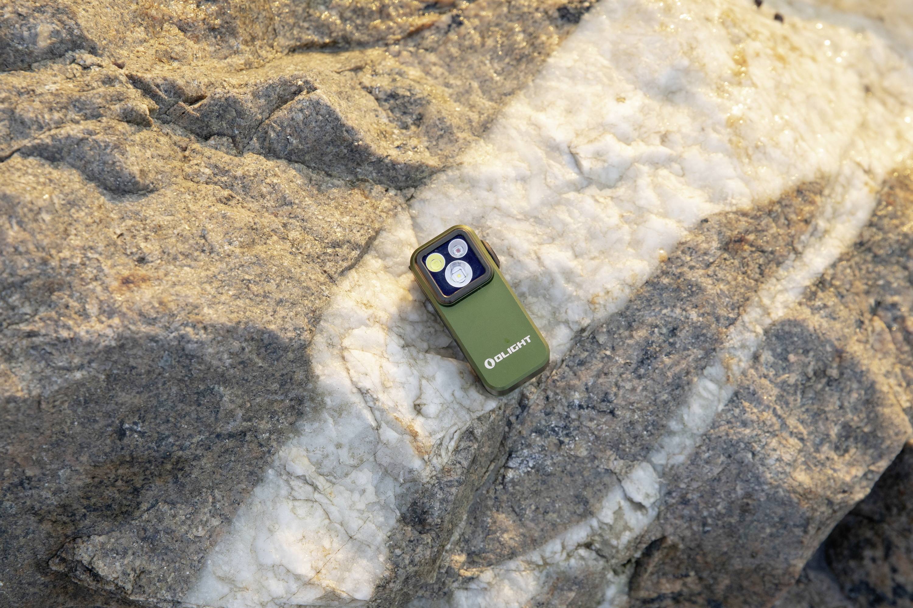 Eine grüne Taschenlampe liegt auf einem Felsen, im Sonnenlicht. Sie hat zwei schmale Lampen und trägt die Marke 'Olight'.