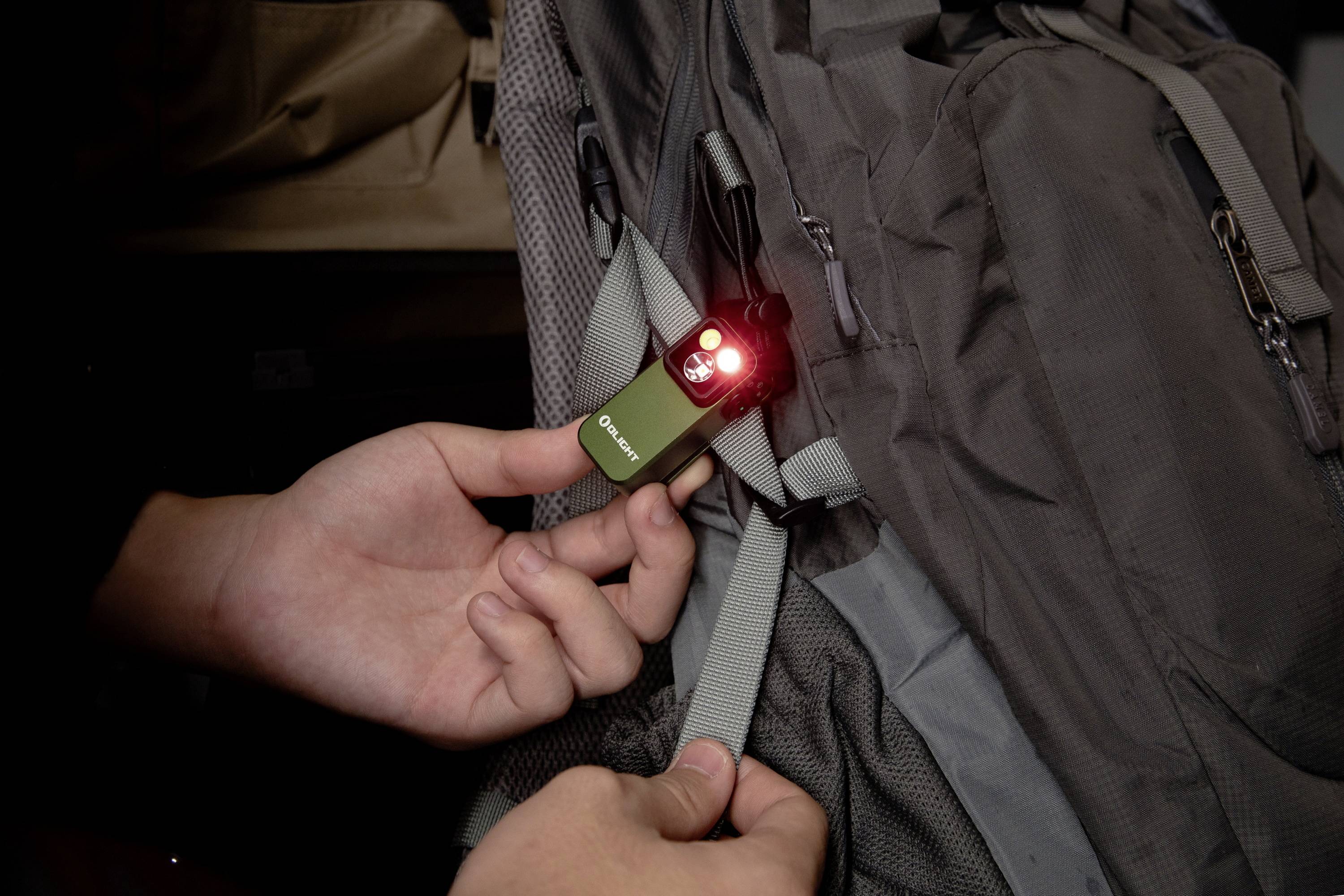 Eine Person befestigt ein kleines, leuchtendes Sicherheitslicht an einem grauen Rucksack, was auf Outdoor-Nutzung hinweist.
