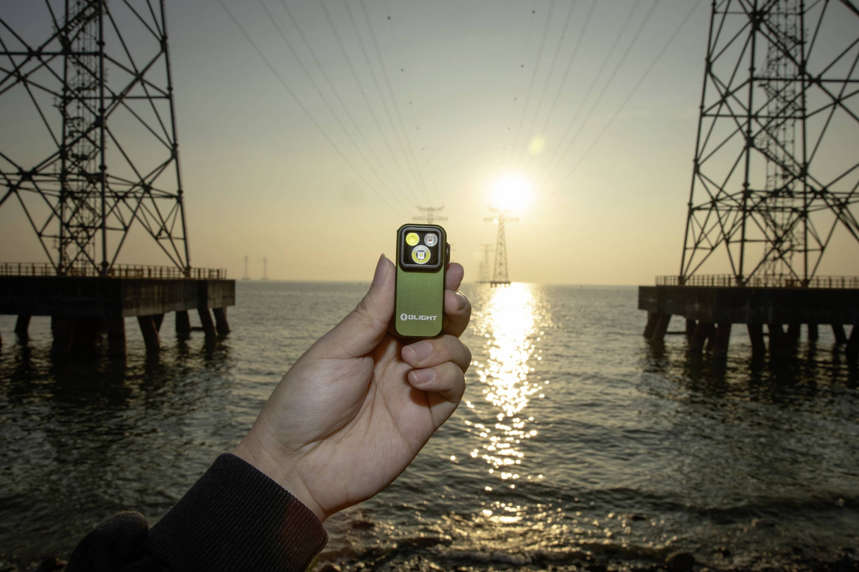 Eine Hand hält eine kleine Taschenlampe vor einem Sonnenuntergang am Meer, zwischen zwei großen Strompylonen.