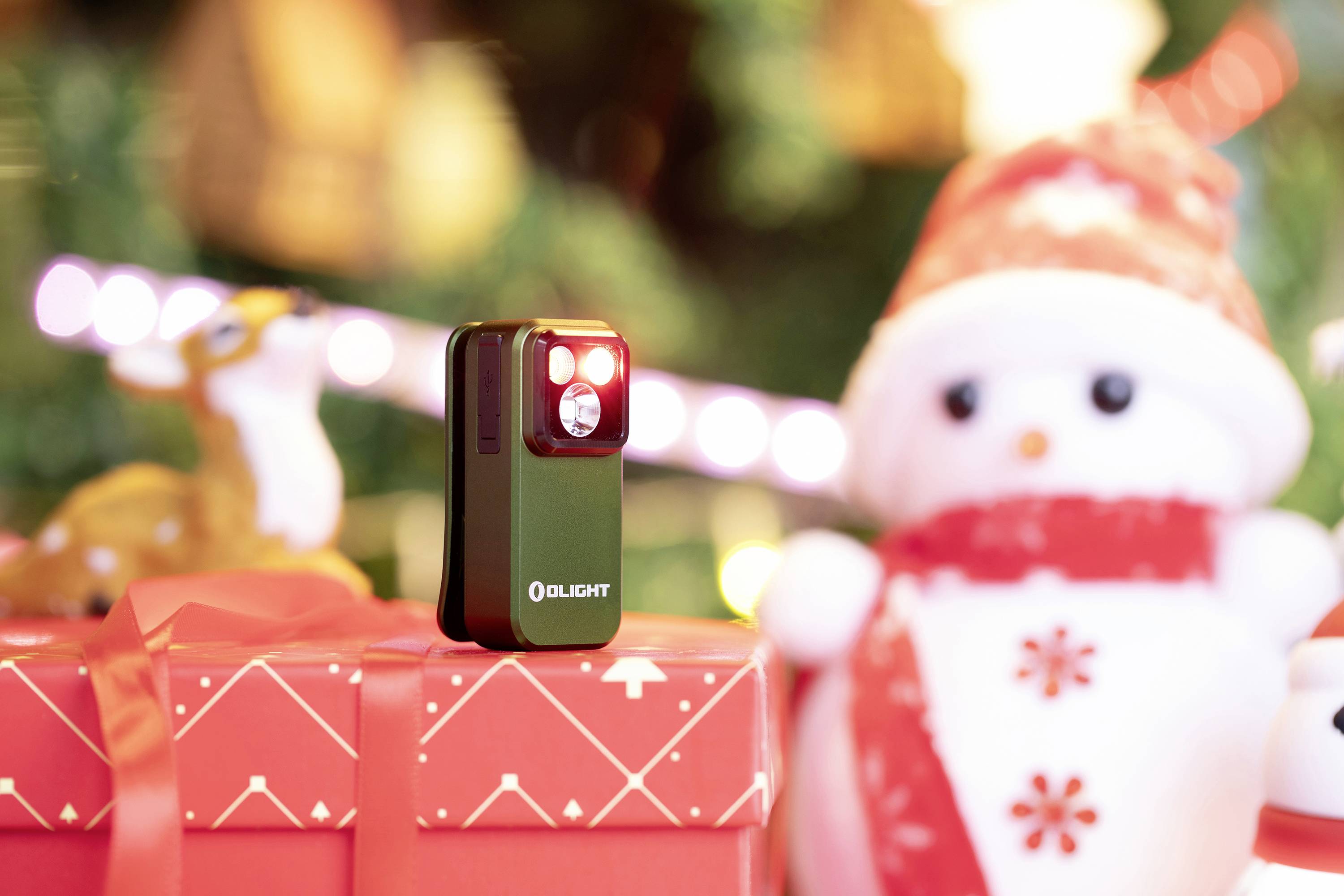 Eine Taschenlampe von Olight steht auf einem rot eingepackten Geschenk, daneben eine Schneemannfigur mit Weihnachtsdeko im Hintergrund.