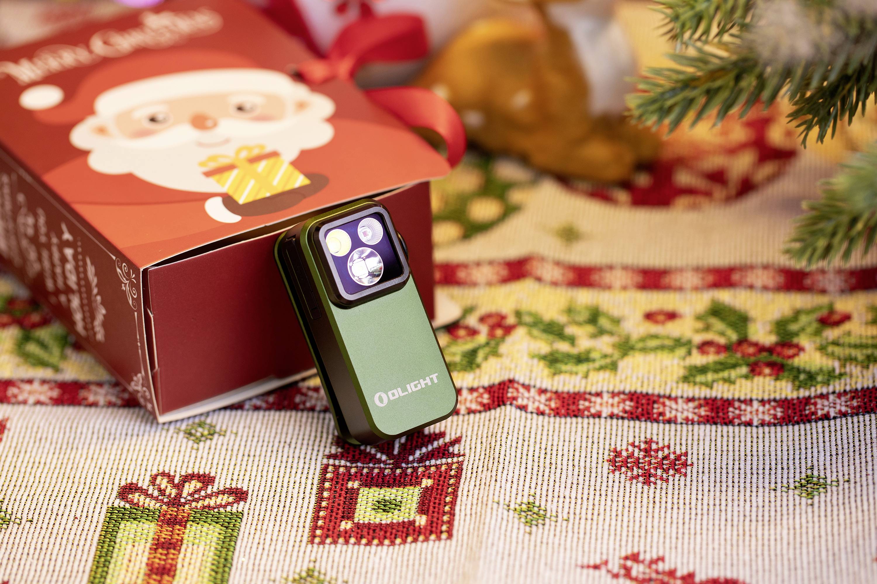 Ein grünes LED-Licht steht neben einer rot-weißen Geschenkbox mit Weihnachtsmotiven auf einem weihnachtlich geschmückten Tisch.