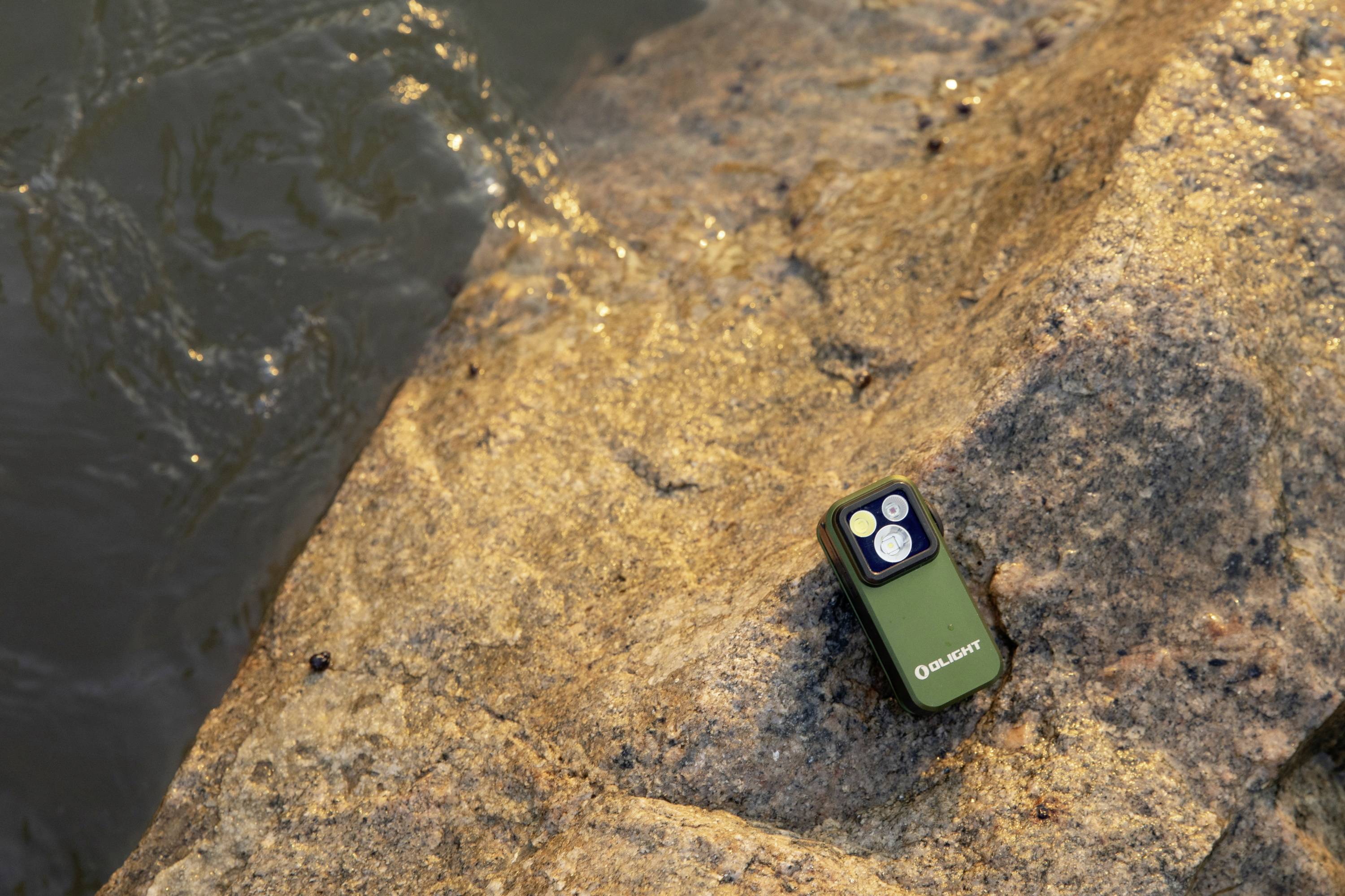 Ein kleines grünes Gerät mit vier LEDs liegt auf einem Felsen am Ufer eines Gewässers. Es ist teilweise beleuchtet von der Sonne.