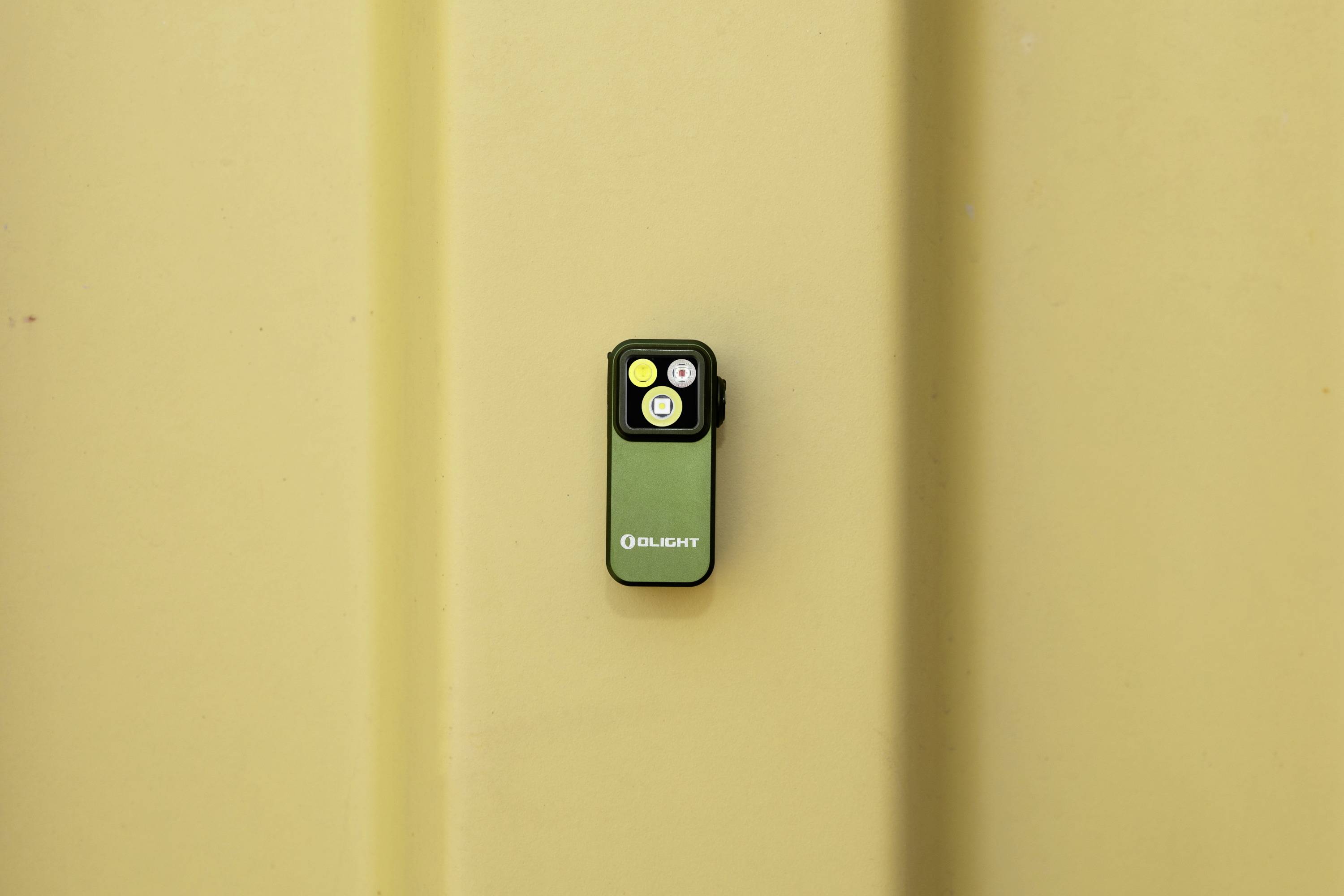 Eine kleine, grüne Taschenlampe ist an einer beigen Wand montiert. Sie hat zwei Knöpfe, einen gelben und einen weißen.