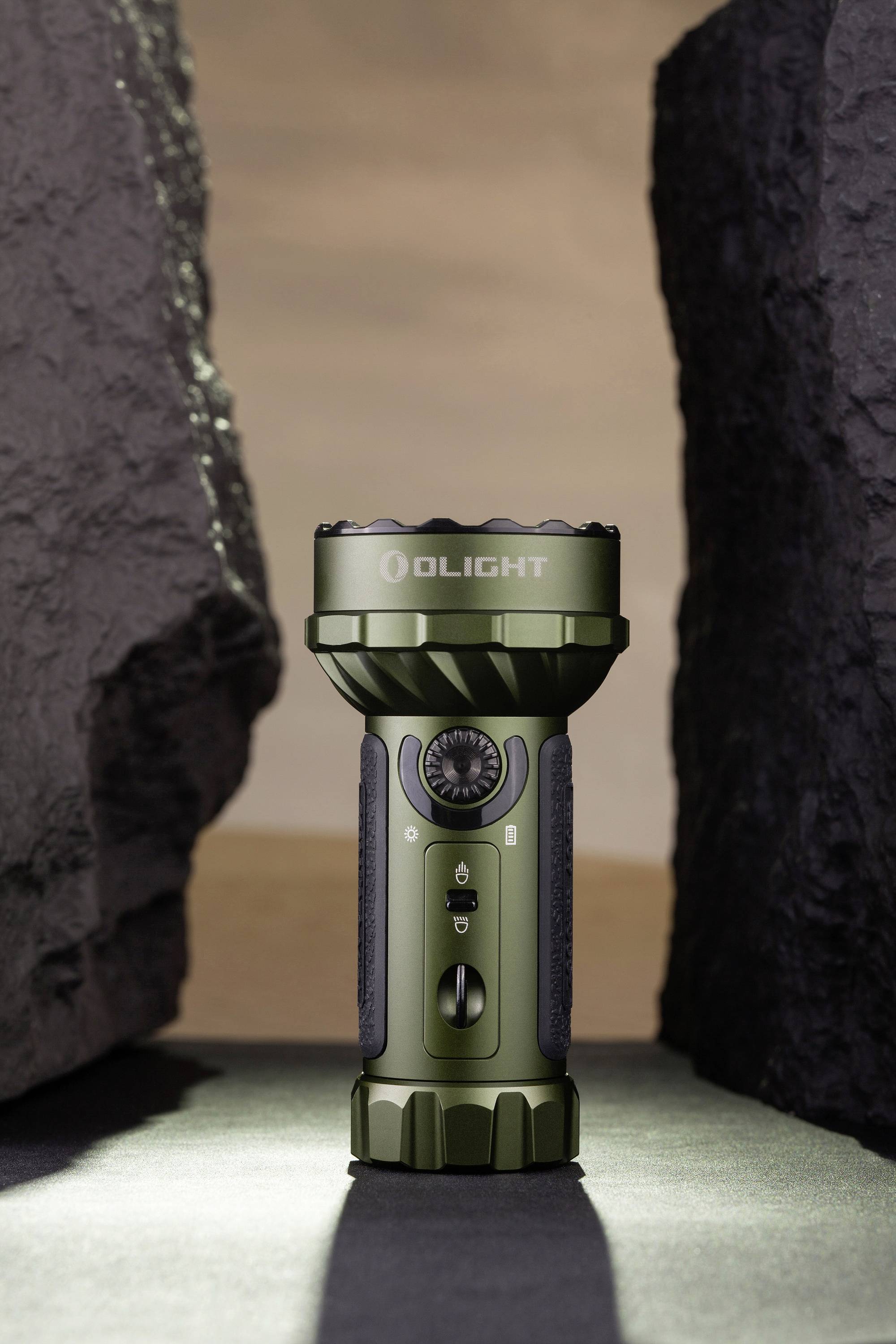 Taschenlampe in olivgrün steht aufrecht zwischen zwei dunklen Felsen vor einem sandfarbenen Hintergrund. Marke 'Olight' sichtbar.