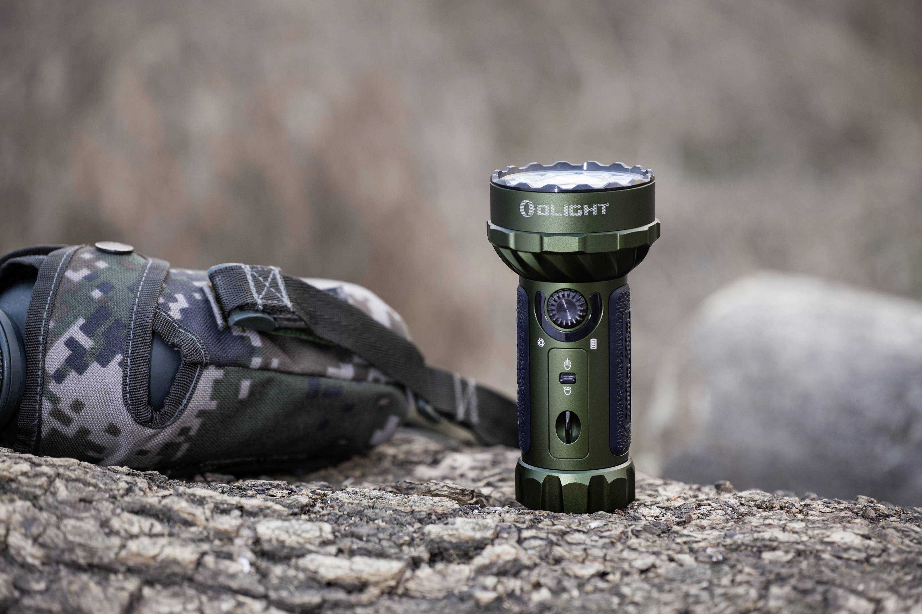Taschenlampe in Tarnfarben liegt auf einem Baumstamm neben einer Tarnstofftasche in einer natürlichen Umgebung.