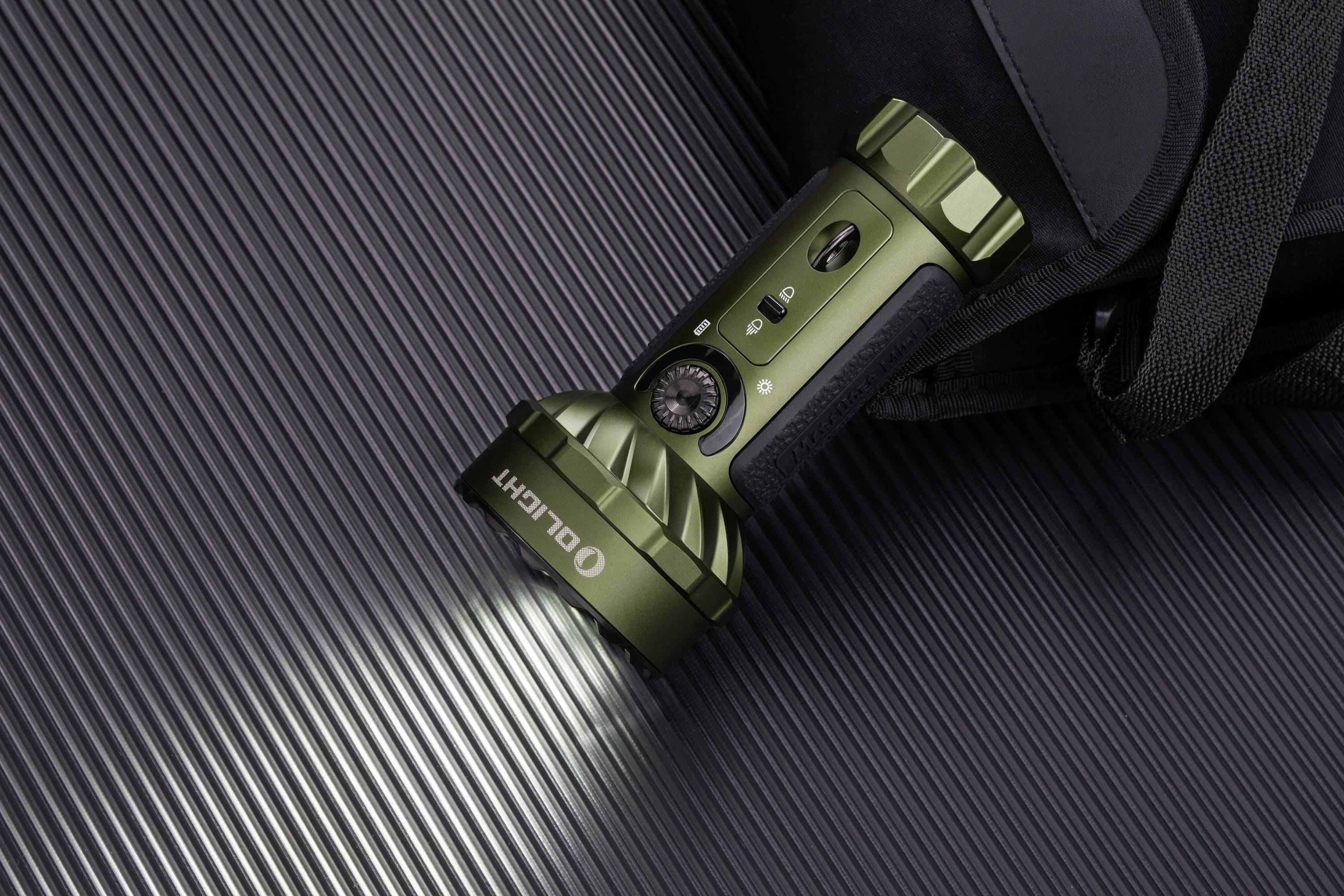 Eine taktische Taschenlampe leuchtet auf eine geriffelte Oberfläche. Sie ist grün, robust und liegt neben einem schwarzen Rucksack.