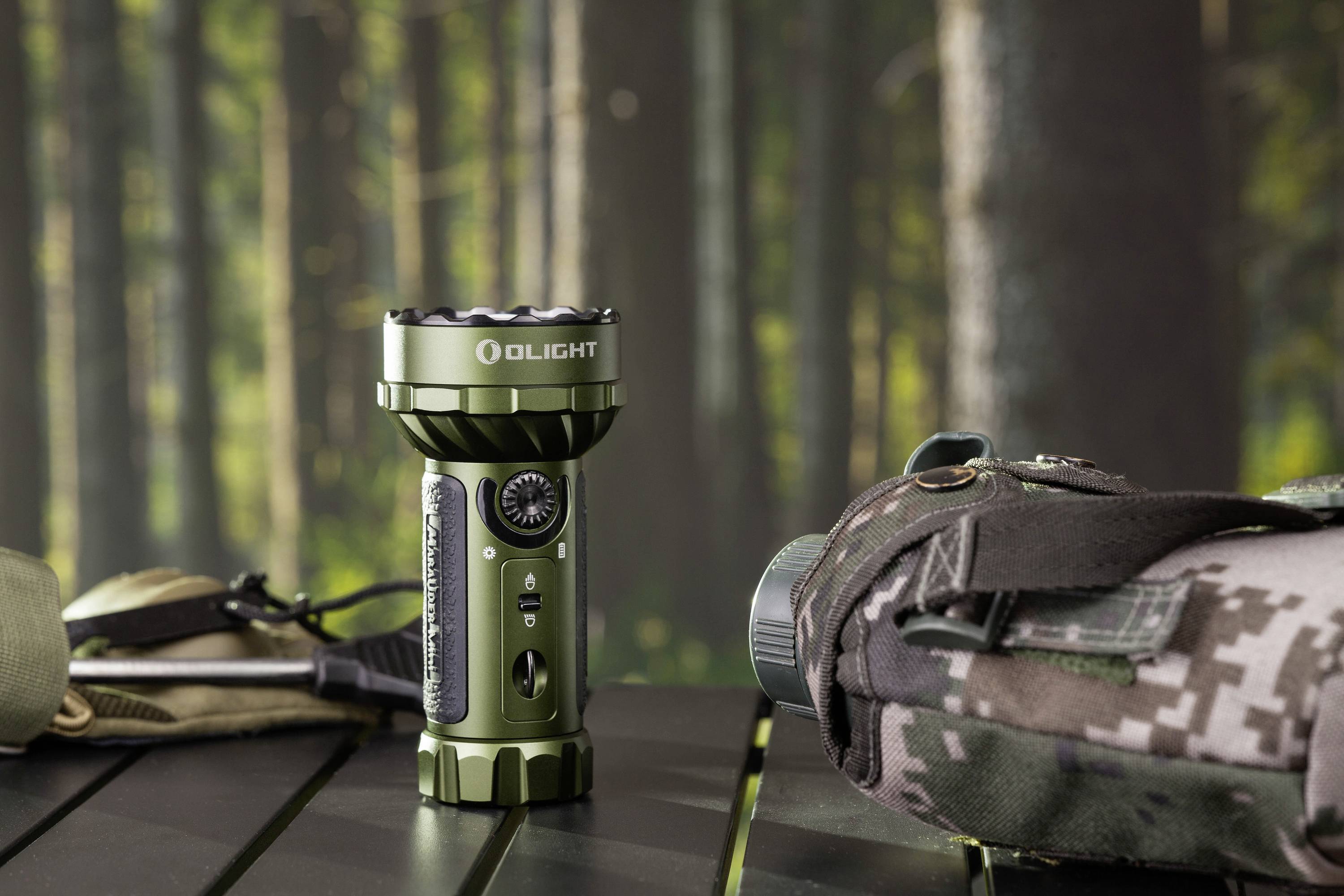 Taschenlampe im Wald auf einem Tisch neben einer Tarnrucksack. Hintergrund unscharf; Waldkulisse erkennbar.