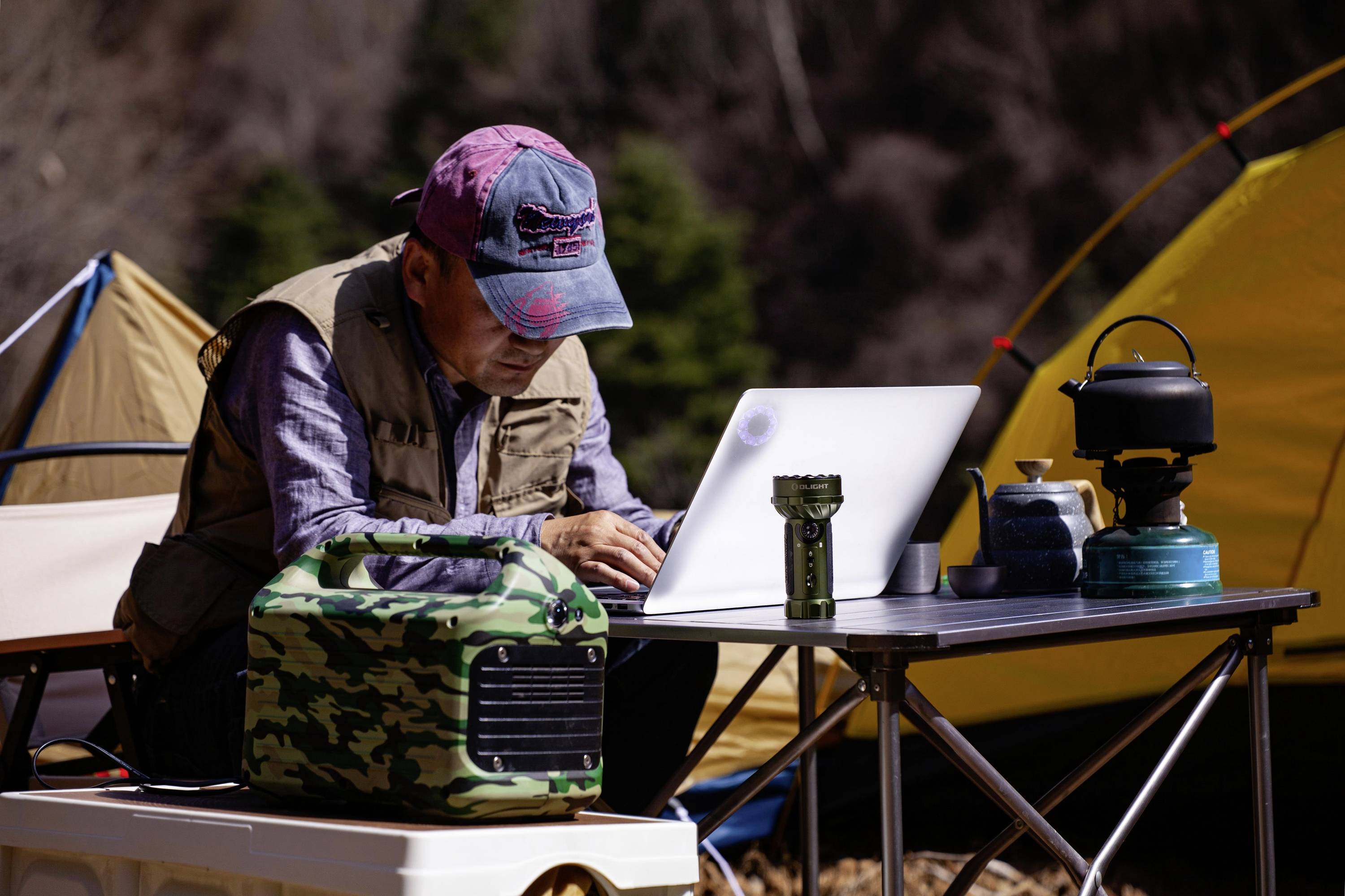 Ein Mann sitzt an einem Campingtisch und arbeitet auf einem Laptop. Um ihn herum sind Campingausrüstung wie Zelt, Lampe und Kocher.