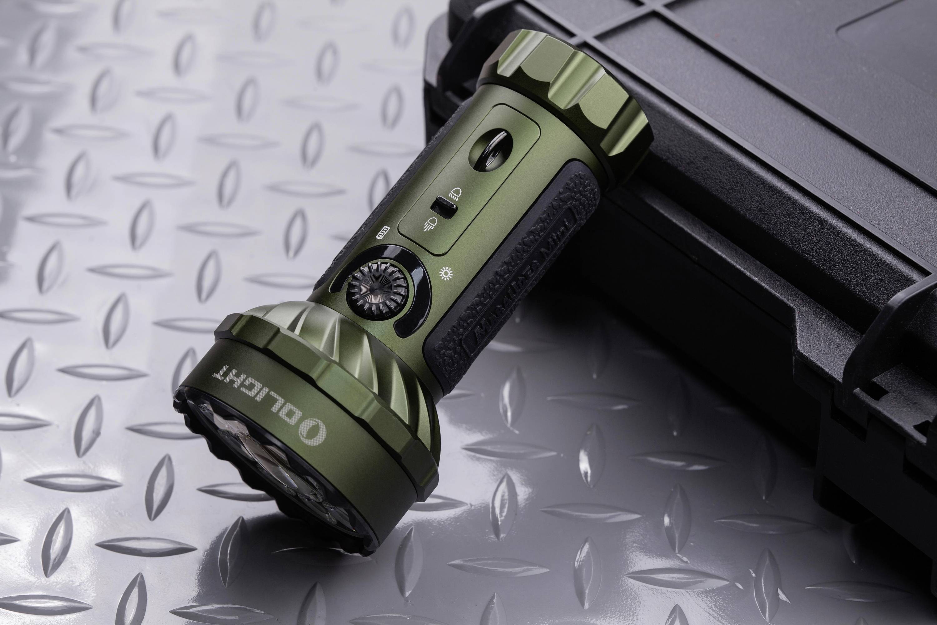 Eine olivgrüne Taschenlampe liegt auf einem metallischen Rautenmusterboden neben einem schwarzen Koffer.