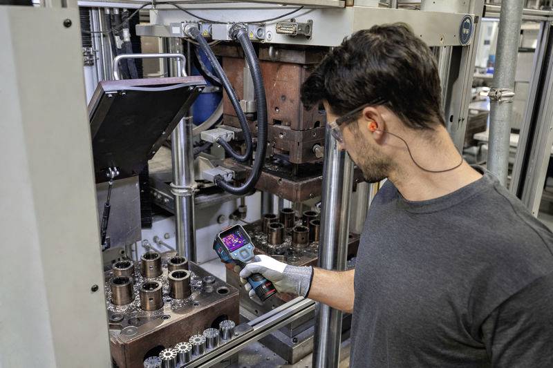 Ein Mann mit Gehörschutz und Handschuhen überwacht Maschinen mit einem Handgerät in einer industriellen Umgebung.