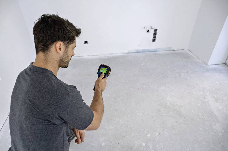 Ein Mann in einem leeren Raum verwendet eine Wärmebildkamera, um die Temperaturverteilung auf dem Boden zu überprüfen.