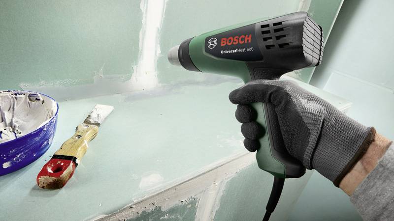'Ein Handschuh hält ein Bosch-Heißluftgebläse nahe einer Wand, wo eine Spachtel und Farbe auf einer Palette sichtbar sind.'