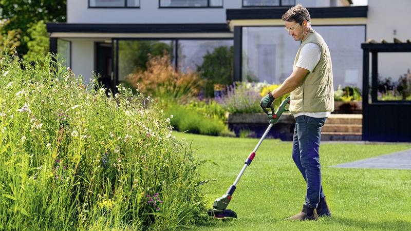 Eine Person mäht mit einem Rasentrimmer den Rasen vor einem modernen Haus. Im Hintergrund ist eine üppige Blumenwiese zu sehen.