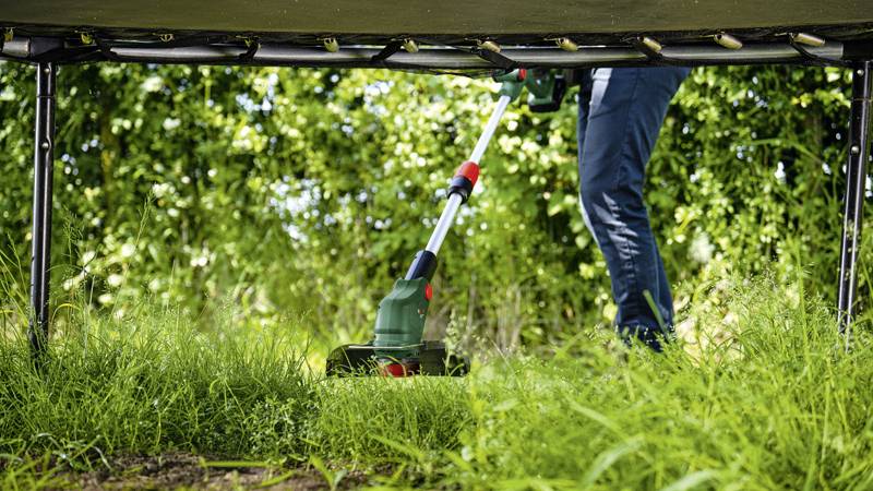 Eine Person hält einen Rasentrimmer und schneidet Gras unter einem Trampolin. Im Hintergrund sind grüne Büsche zu sehen.