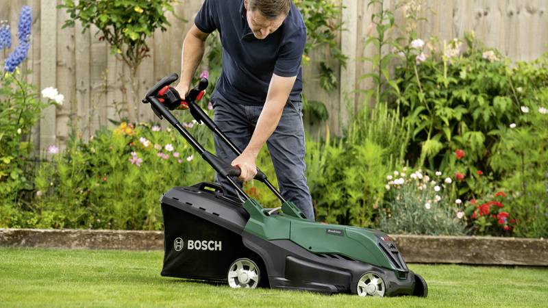Eine Person mäht den Rasen mit einem grünen Bosch-Rasenmäher im Garten, umgeben von blühenden Blumenbeeten und einem Holzzaun.