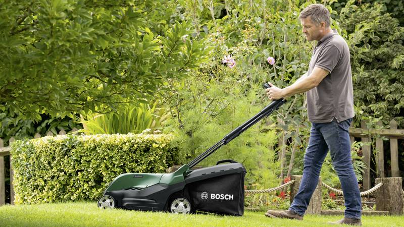 Ein Mann mäht den Rasen mit einem grünen Bosch-Rasenmäher in einem gepflegten Garten, umgeben von Büschen und Blumen.