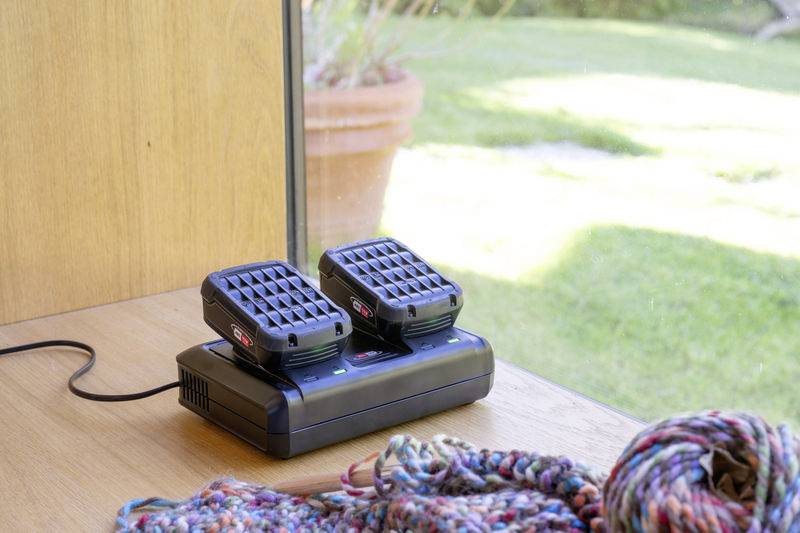Zwei Akkuladegeräte auf einem Tisch vor einem Fenster mit Blick auf einen Garten. Im Vordergrund liegt ein bunter Schal.