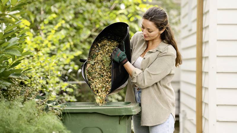 Eine Frau leert einen Behälter mit Gartenabfällen in eine grüne Tonne, umgeben von Pflanzen und einem Gebäude im Hintergrund.
