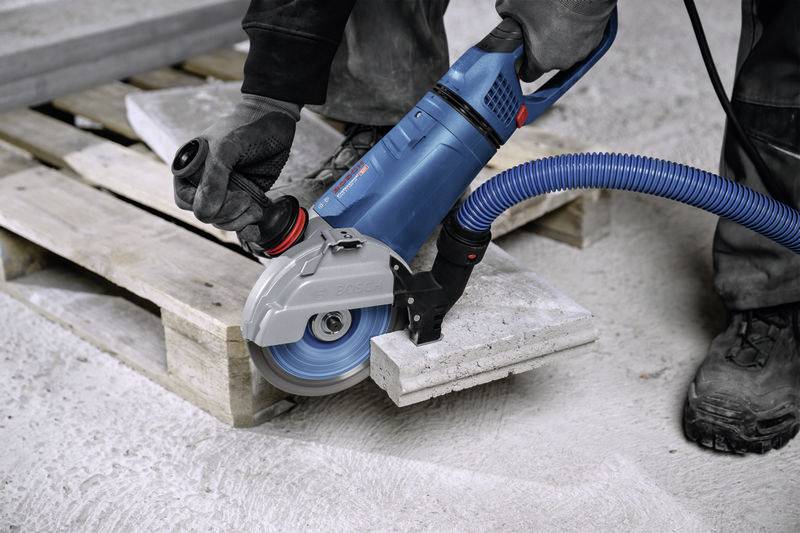 Eine Person schneidet mit einer elektrischen Schleifmaschine einen Betonblock auf einer Palette. Staub wird durch einen blauen Schlauch abgesaugt.