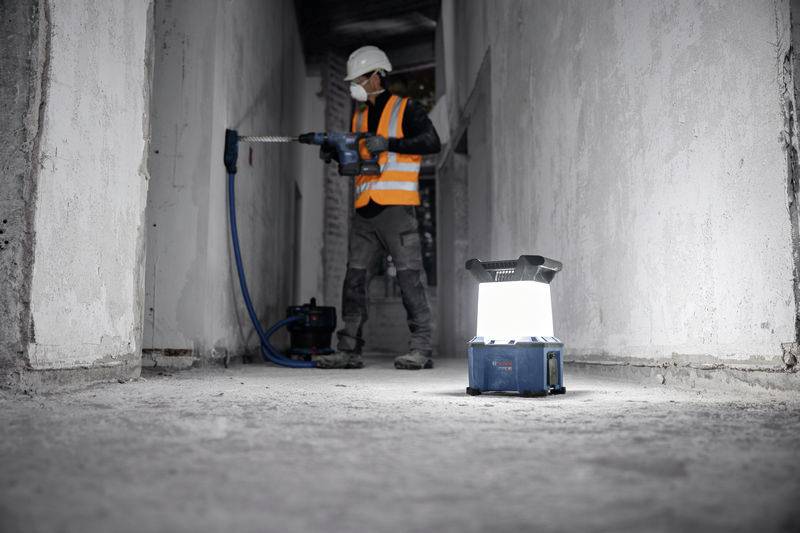 Ein Bauarbeiter mit Sicherheitsweste und Helm bohrt in eine Wand in einem verlassenen Gebäude. Eine leuchtende Bauleuchte steht im Vordergrund.