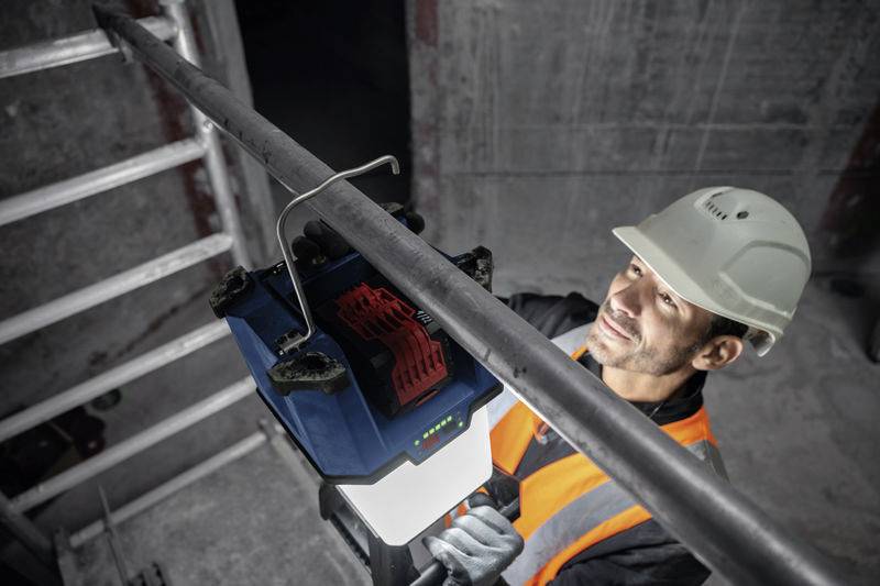 Ein Bauarbeiter mit Helm und orangefarbener Weste benutzt ein Vermessungsgerät. Er arbeitet neben einer Leiter in einem Betonraum.