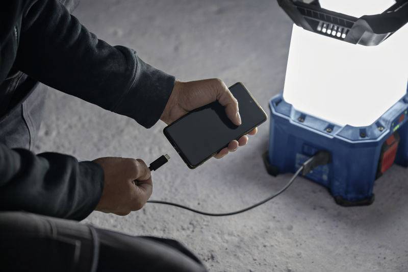 Eine Person lädt ein Smartphone an einer tragbaren LED-Laterne auf, die auf einem Betonboden steht.