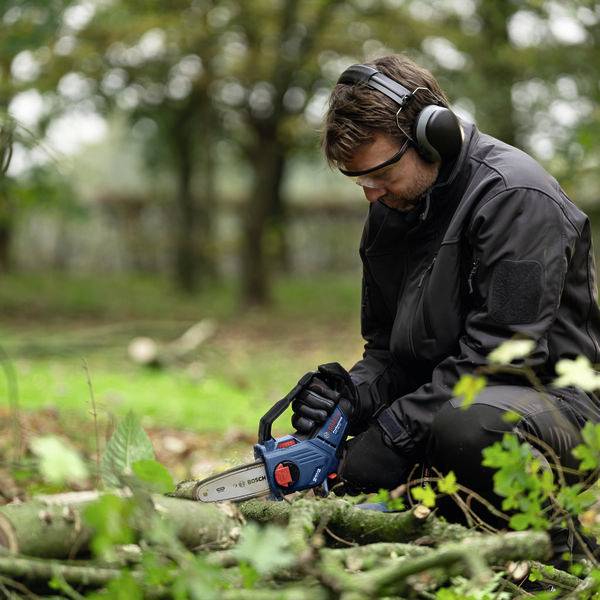 Eine Person in Schutzkleidung sägt mit einer Kettensäge Holz auf einer grünen Wiese. Im Hintergrund stehen Bäume.