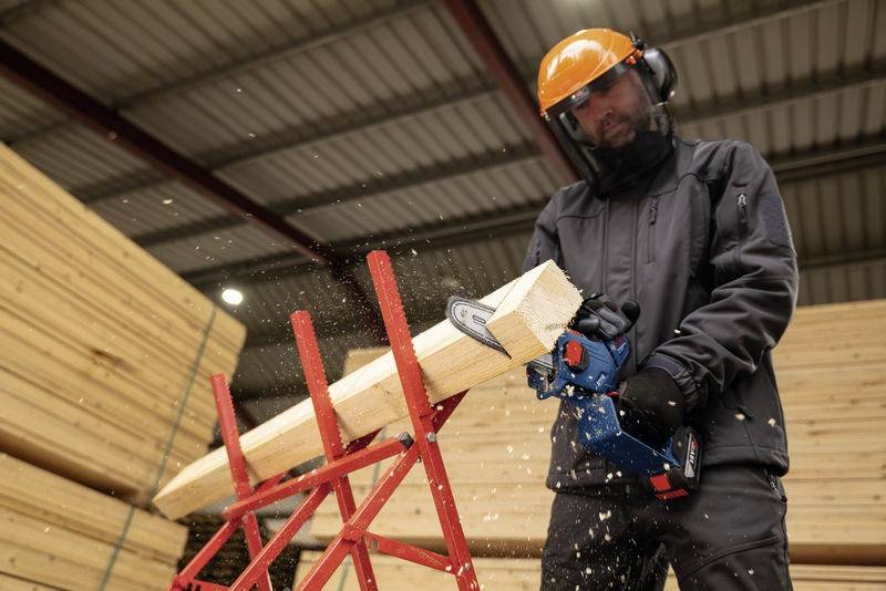 Eine Person mit Schutzhelm und Brille schneidet mit einer elektrischen Säge einen Holzblock in einem Lagerhaus voller Holzstapel.