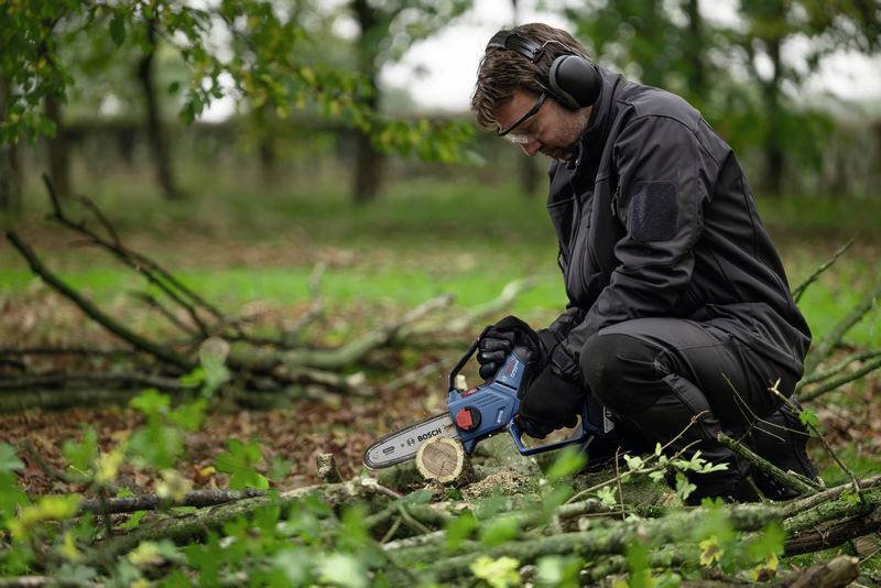 Eine Person sägt mit einer elektrischen Kettensäge Holz auf einem baumbestandenen Gelände. Sie trägt Schutzkleidung und Gehörschutz.