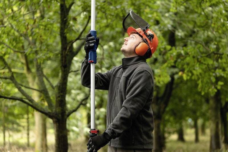 Ein Mann mit Schutzhelm und Gehörschutz bedient eine Teleskopstange in einem Wald, während er nach oben schaut.